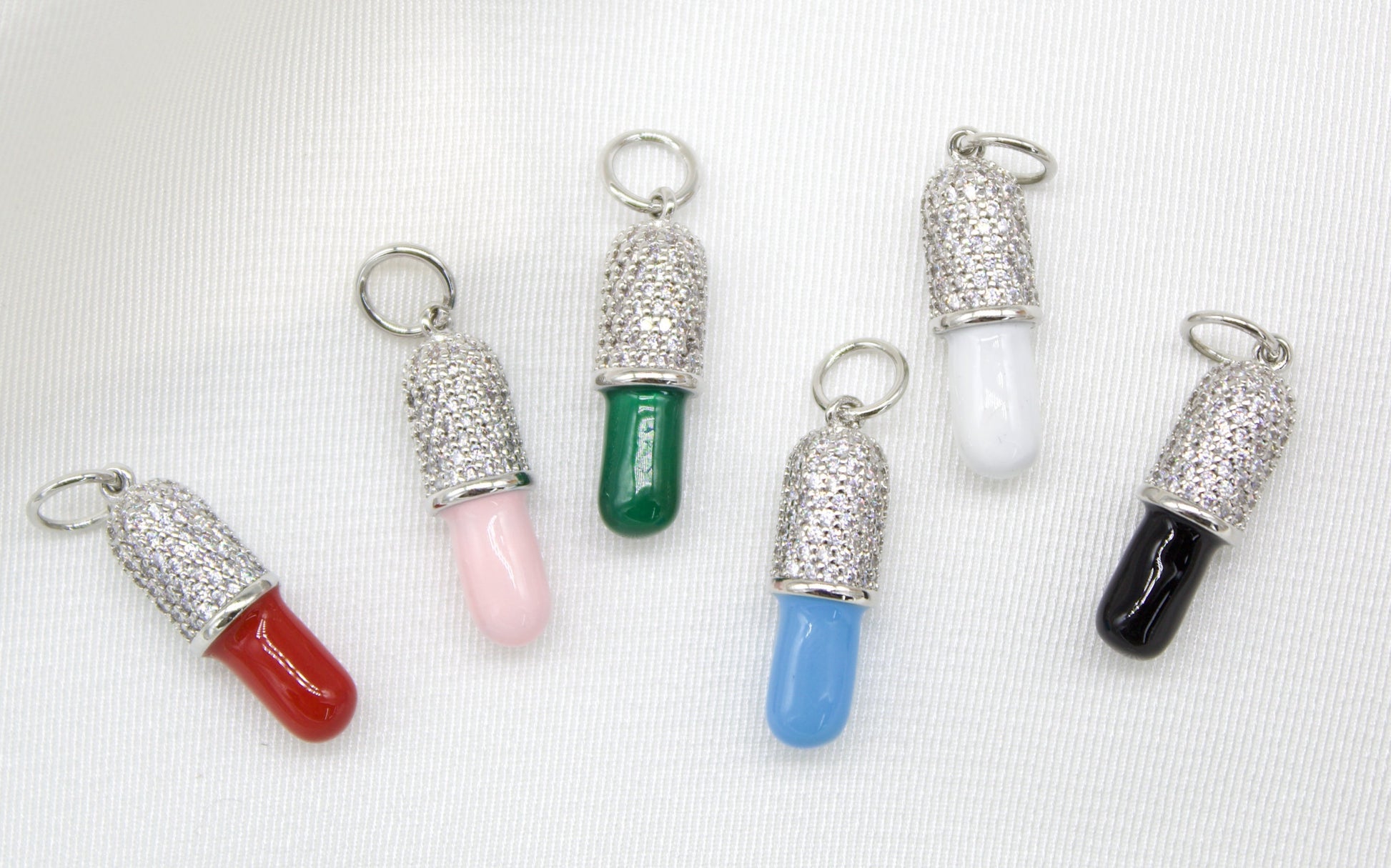 Six colorful pill-shaped pendant charms featuring paved cubic zirconia crystals and enamel details rest on a white fabric background.