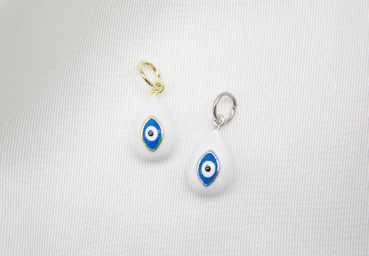 Two teardrop-shaped enamel evil eye charms, one in gold and one in silver, rest on a white fabric background.