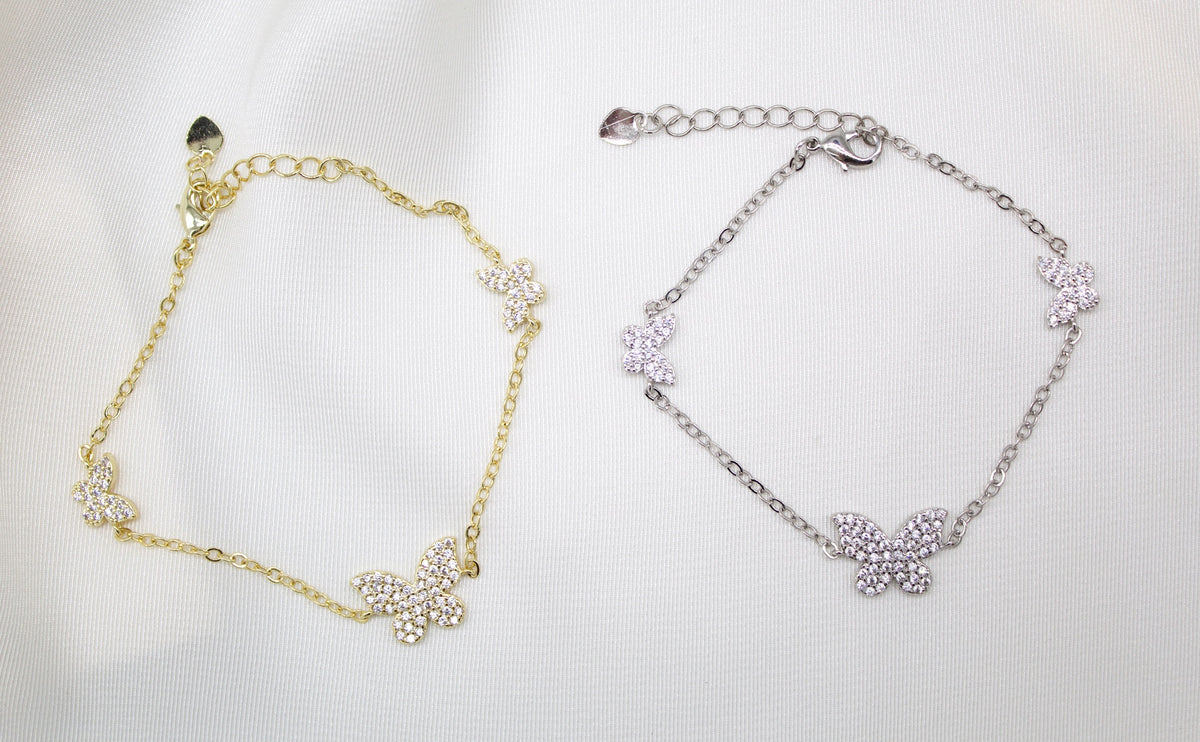 A gold and a silver butterfly charm bracelet rest side by side on a white fabric background, each sparkling with inlaid cubic zirconia crystals.