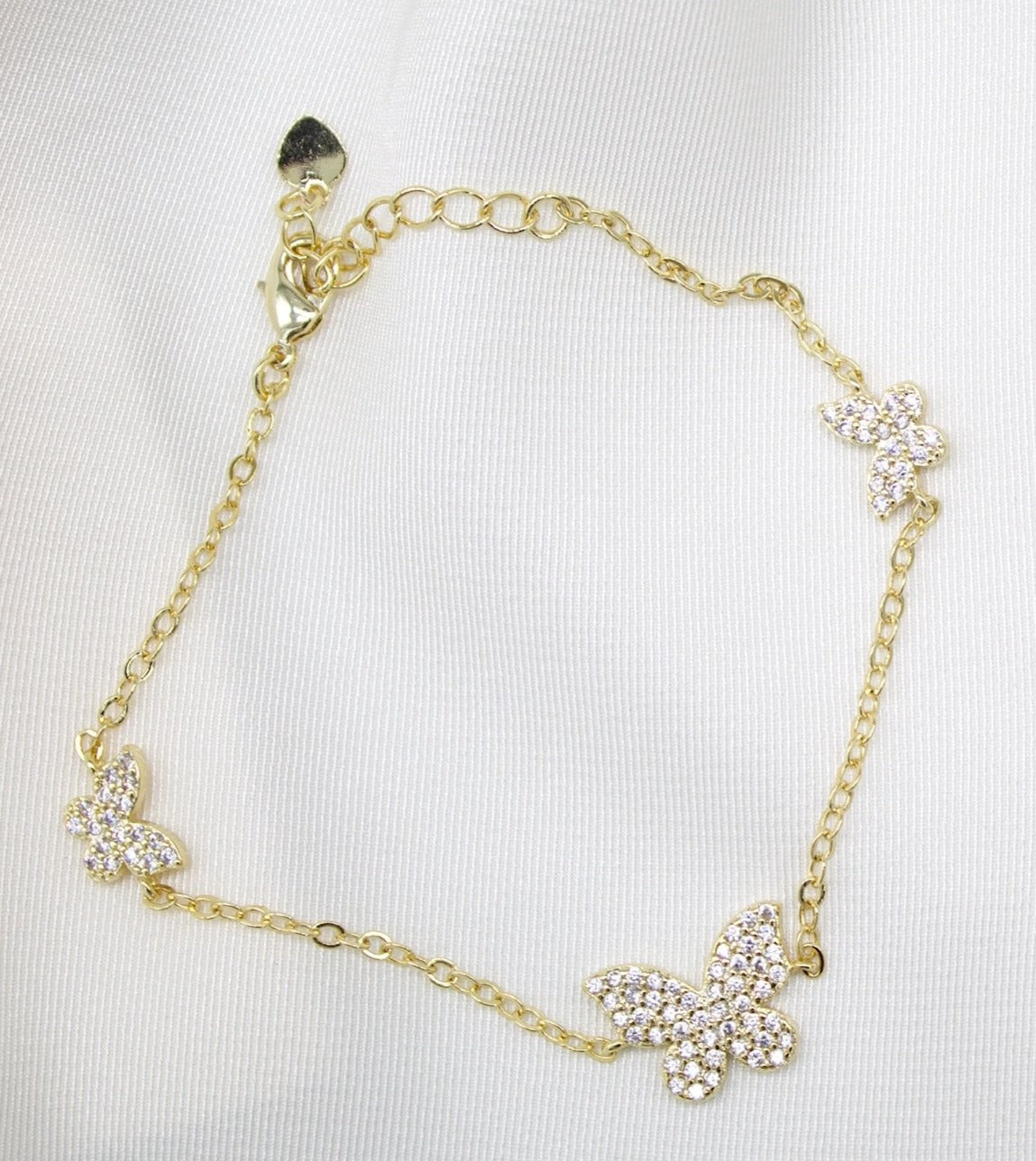 A delicate gold chain bracelet features three pavé crystal-encrusted butterfly charms resting against a white fabric background.