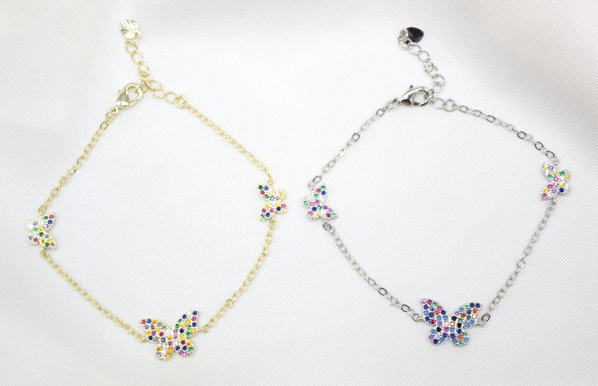 Two delicate butterfly charm bracelets, one gold and one silver, display colorful gemstone accents against a white satin background.