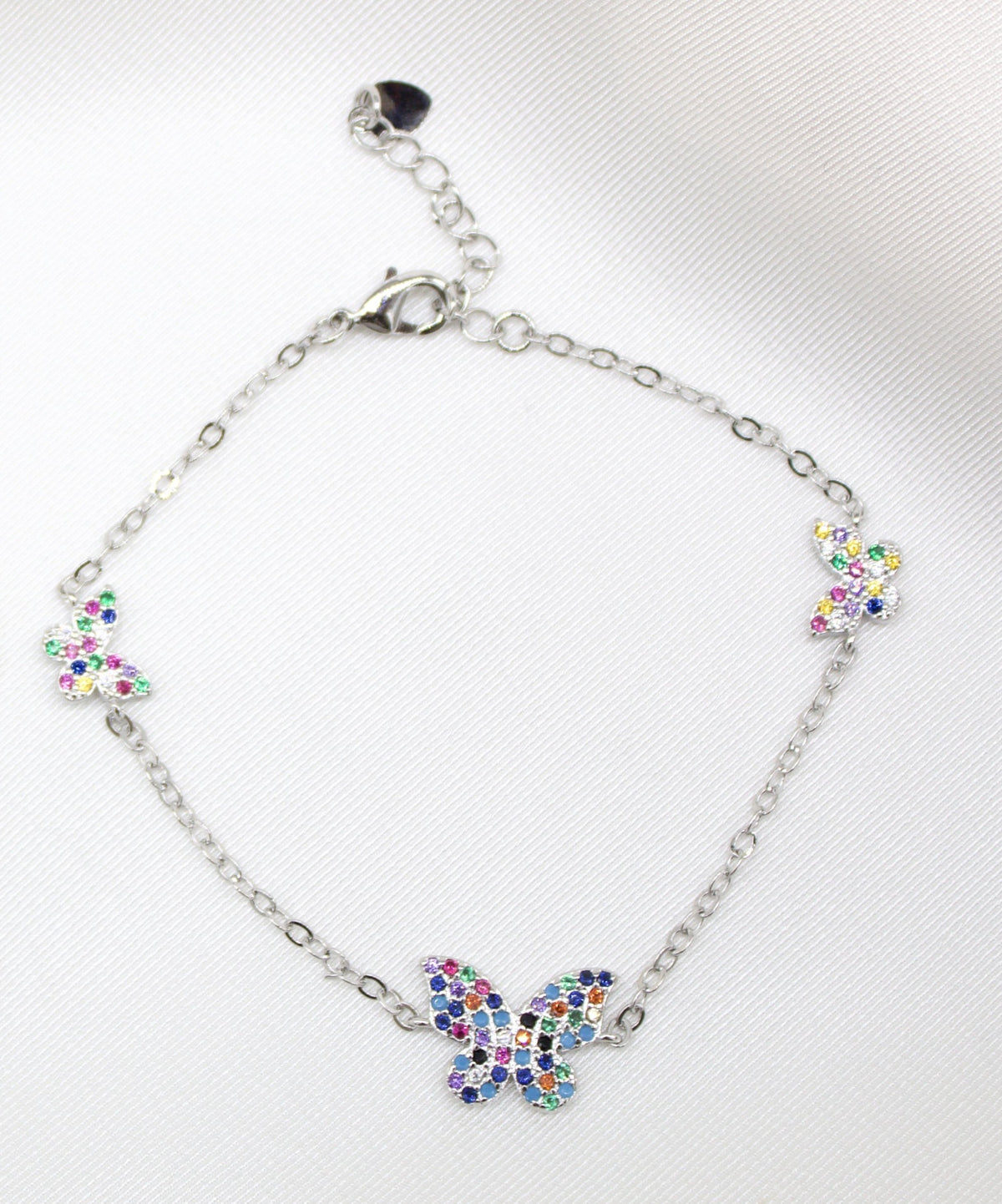 A delicate silver chain bracelet features three colorful, crystal-studded butterfly charms against a white satin background.