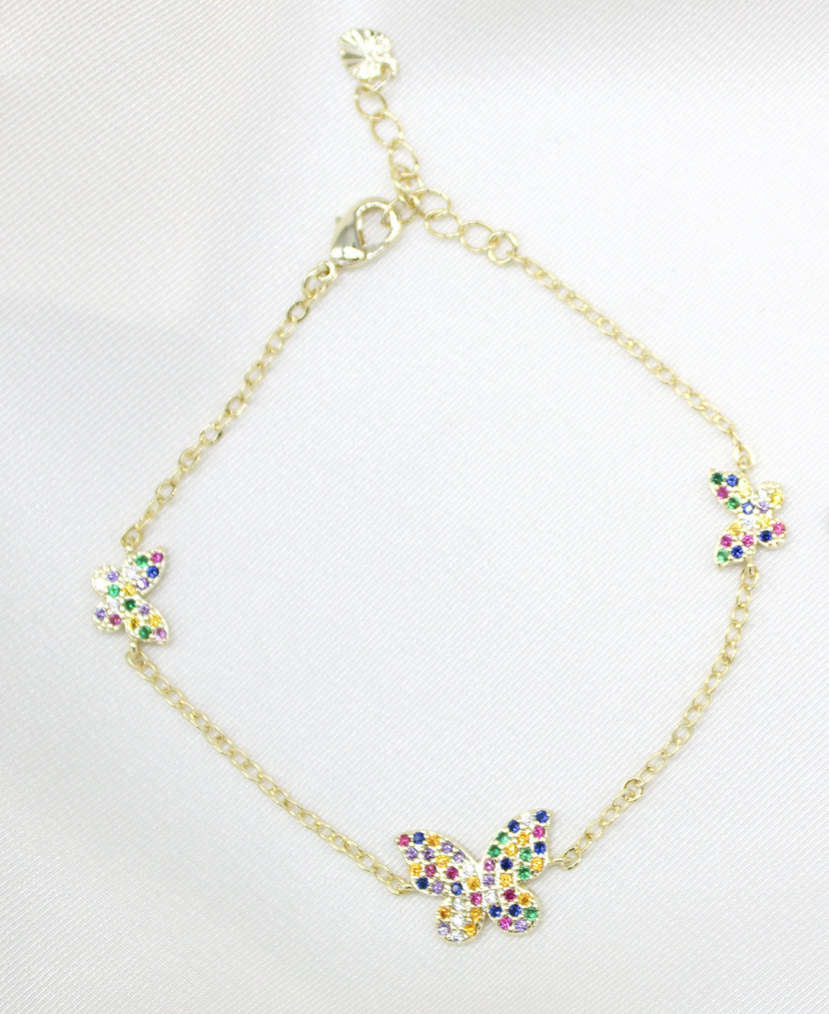 A delicate gold chain bracelet features three colorful, crystal-encrusted butterfly charms displayed on a white satin background.
