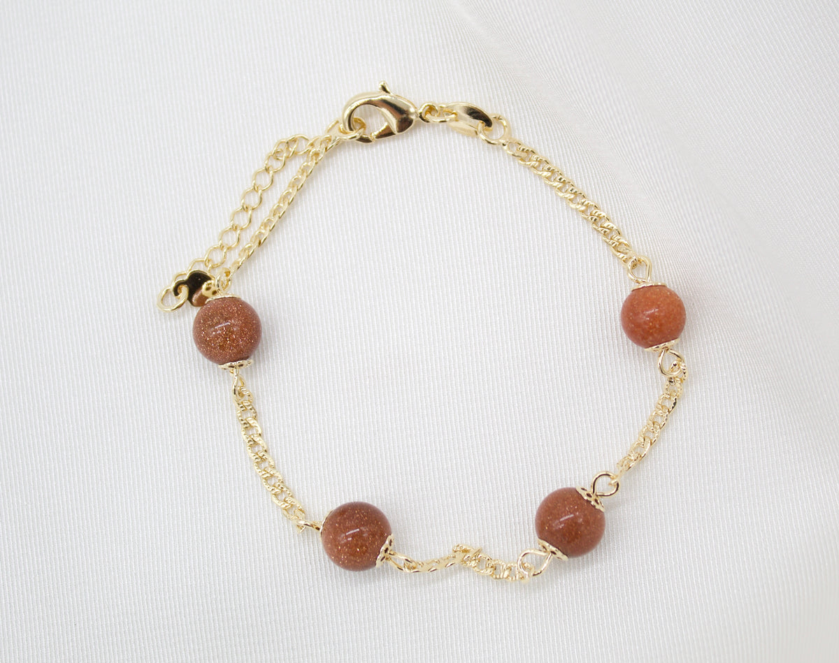 A delicate gold-plated chain bracelet featuring sparkling brown sandstone beads rests on a white satin background.