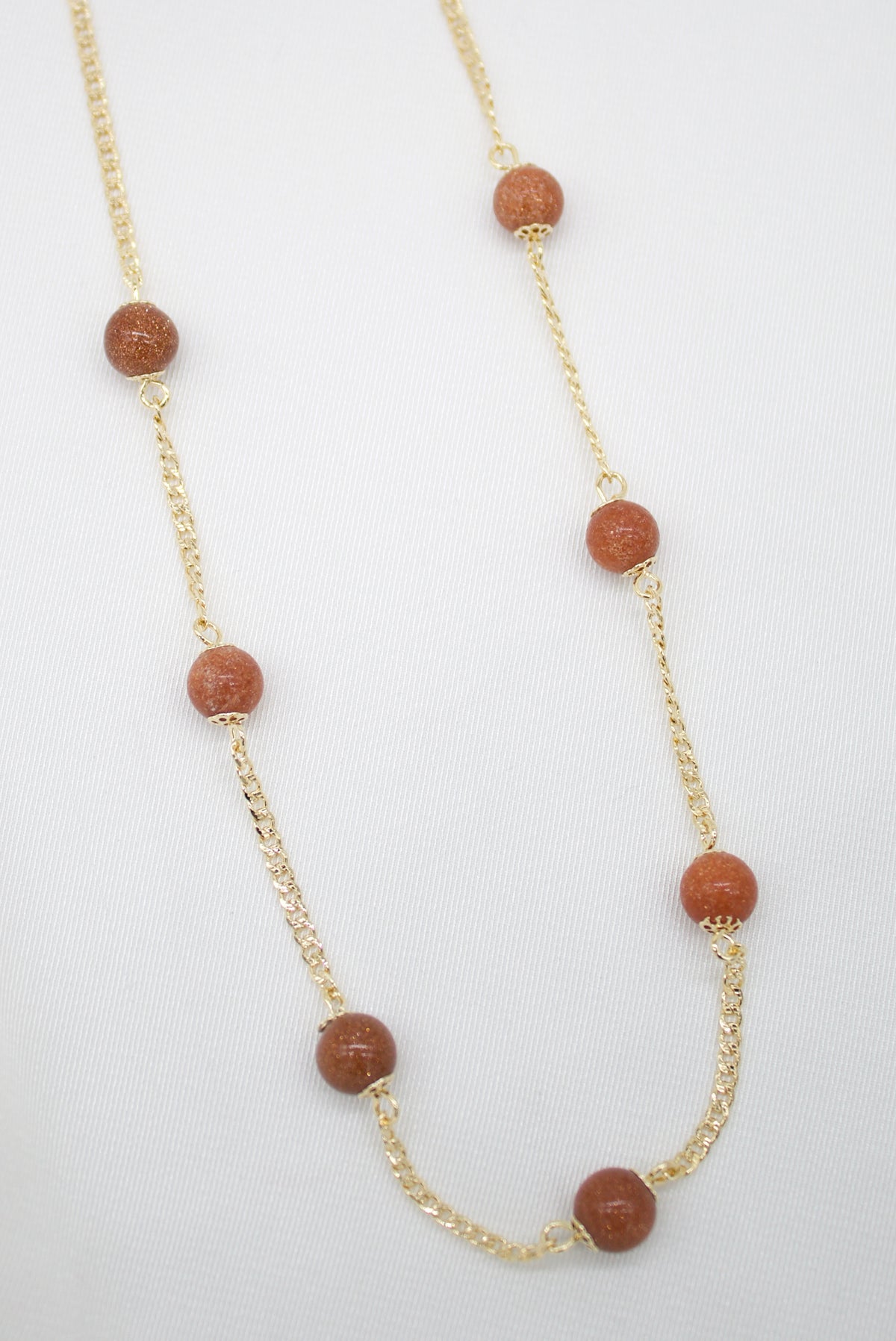 A delicate gold chain necklace features evenly spaced round brown sandstone beads against a white background.