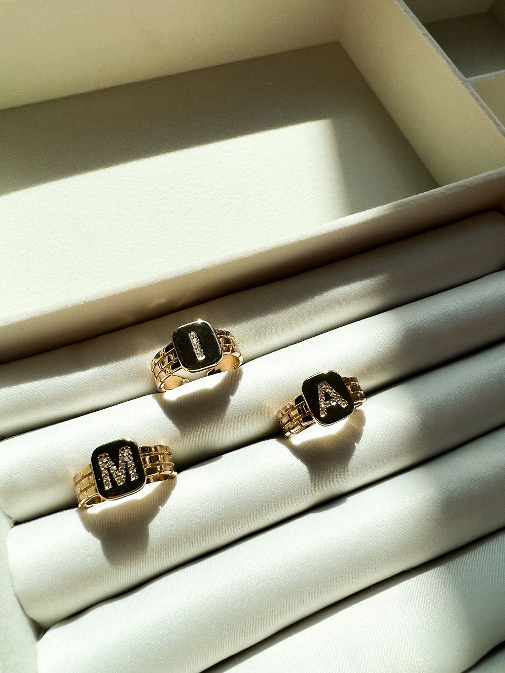 Three gold initial rings spelling "MIA" rest neatly inside a soft white velvet jewelry box.