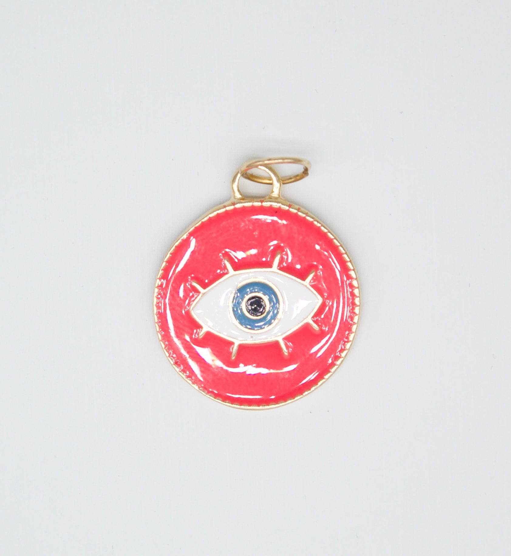 A round gold evil eye pendant with vibrant pink enamel features a white and blue eye symbol at the center.