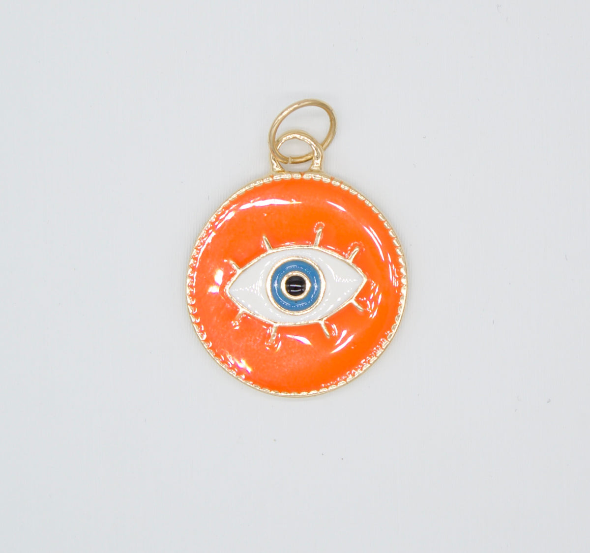 A vibrant orange evil eye pendant featuring an enamel finish and gold-toned borders rests against a plain white background.