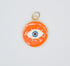 A vibrant orange evil eye pendant featuring an enamel finish and gold-toned borders rests against a plain white background.
