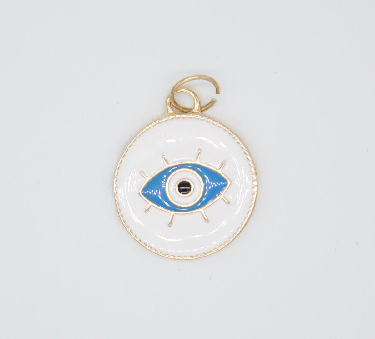 A gold-trimmed white enamel evil eye charm features a vibrant blue and black center against a neutral background.