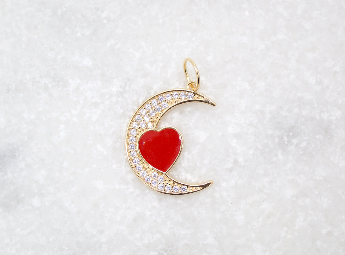 A gold crescent moon pendant rests on a white textured background, featuring sparkling cubic zirconia pavé and a glossy red enamel heart in the center.