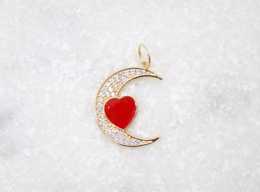 A gold crescent moon pendant rests on a white textured background, featuring sparkling cubic zirconia pavé and a glossy red enamel heart in the center.
