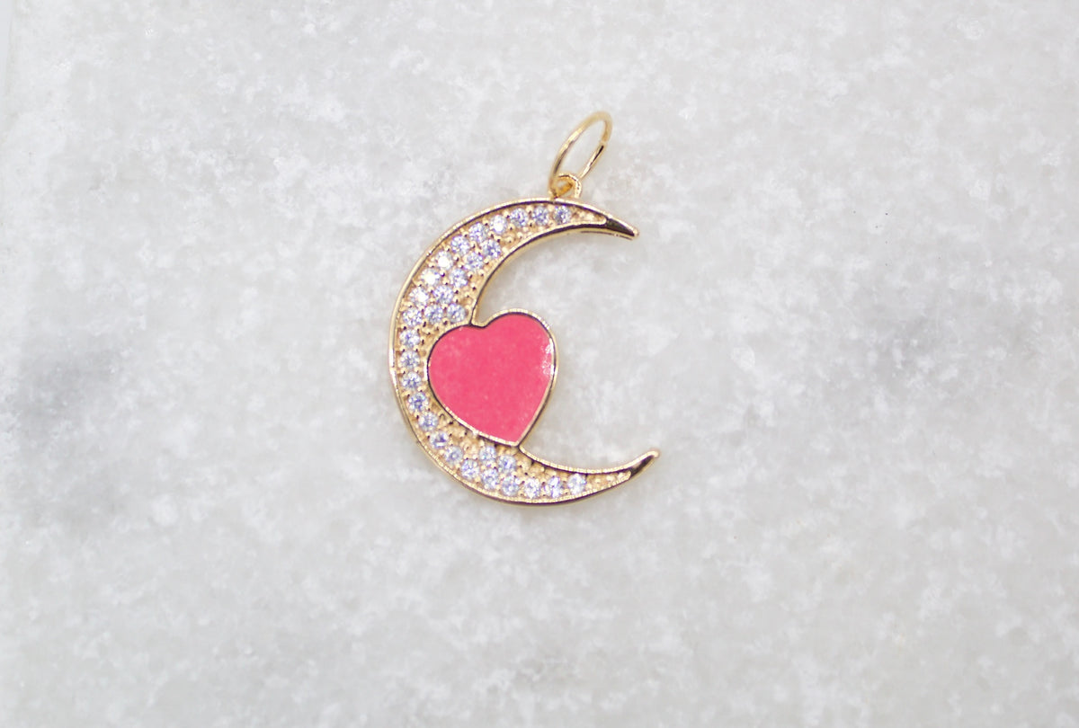 A gold crescent moon pendant rests on a white marble surface, featuring a pink enamel heart and shimmering cubic zirconia stones.