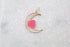 A gold crescent moon pendant rests on a white marble surface, featuring a pink enamel heart and shimmering cubic zirconia stones.