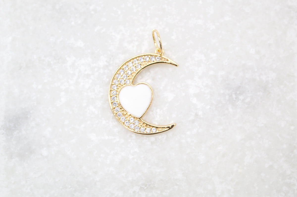 A delicate gold crescent moon pendant sparkles with pavé crystals, featuring a glossy white enamel heart nestled in the center against a textured white marble background.
