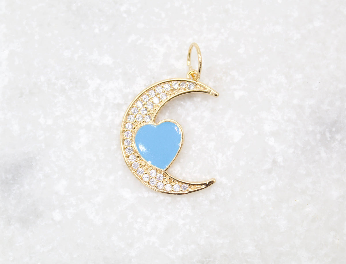 A gold crescent moon pendant rests on a white marble surface, featuring pavé-set CZ stones and a central light blue heart charm.