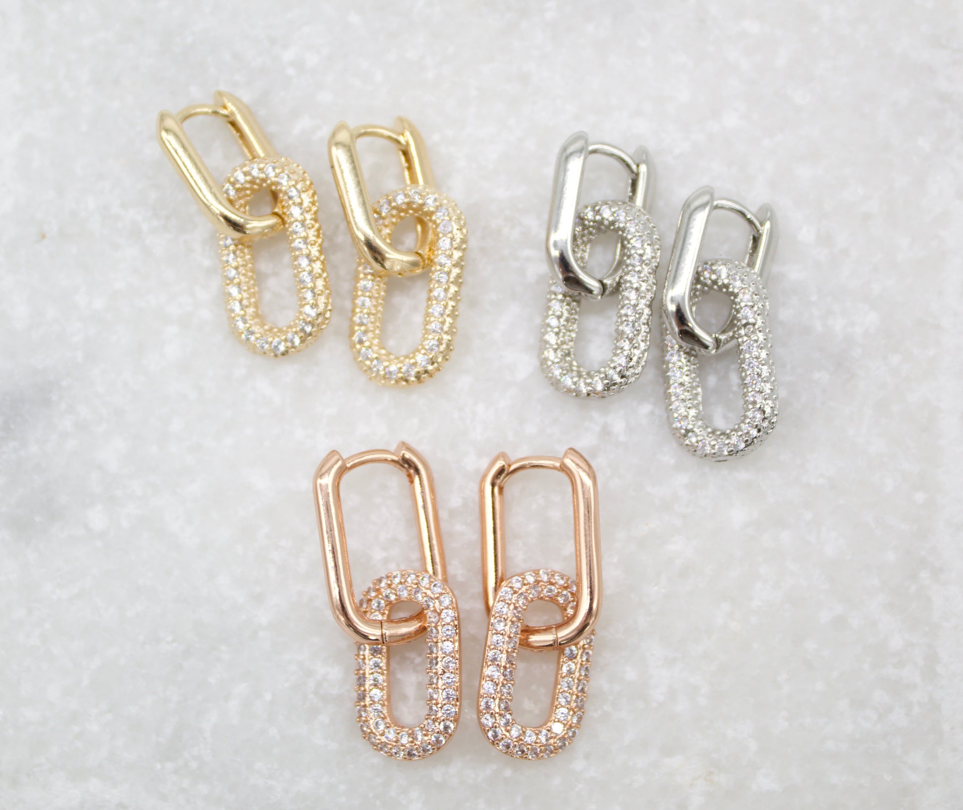 Three pairs of interlocking oval link earrings in gold, silver, and rose gold rest on a white textured surface, showcasing pavé-set cubic zirconia stones.