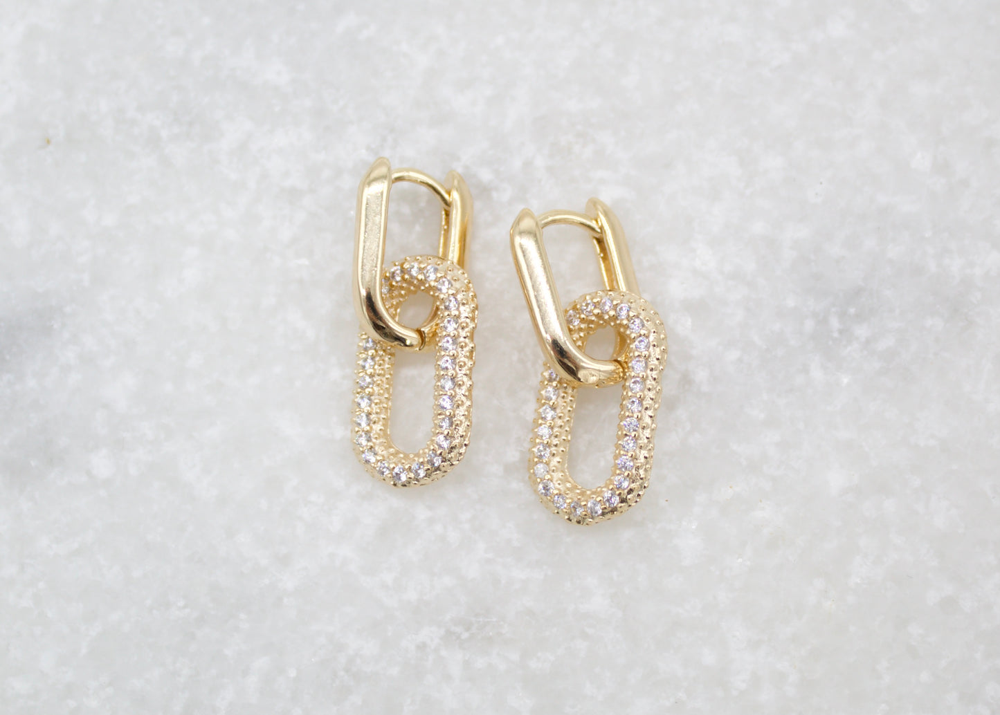A pair of interlocking gold chain-link huggie earrings encrusted with sparkling pave stones rests on a textured white marble surface.
