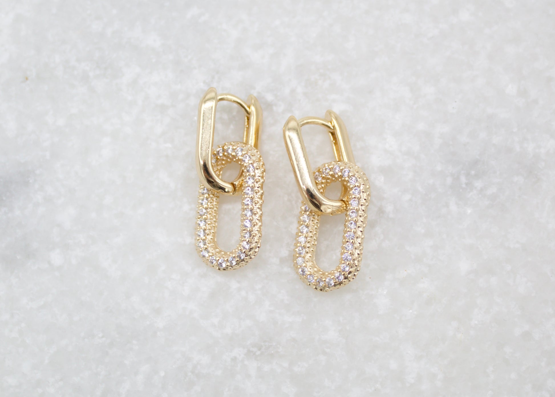 A pair of interlocking gold chain-link huggie earrings encrusted with sparkling pave stones rests on a textured white marble surface.
