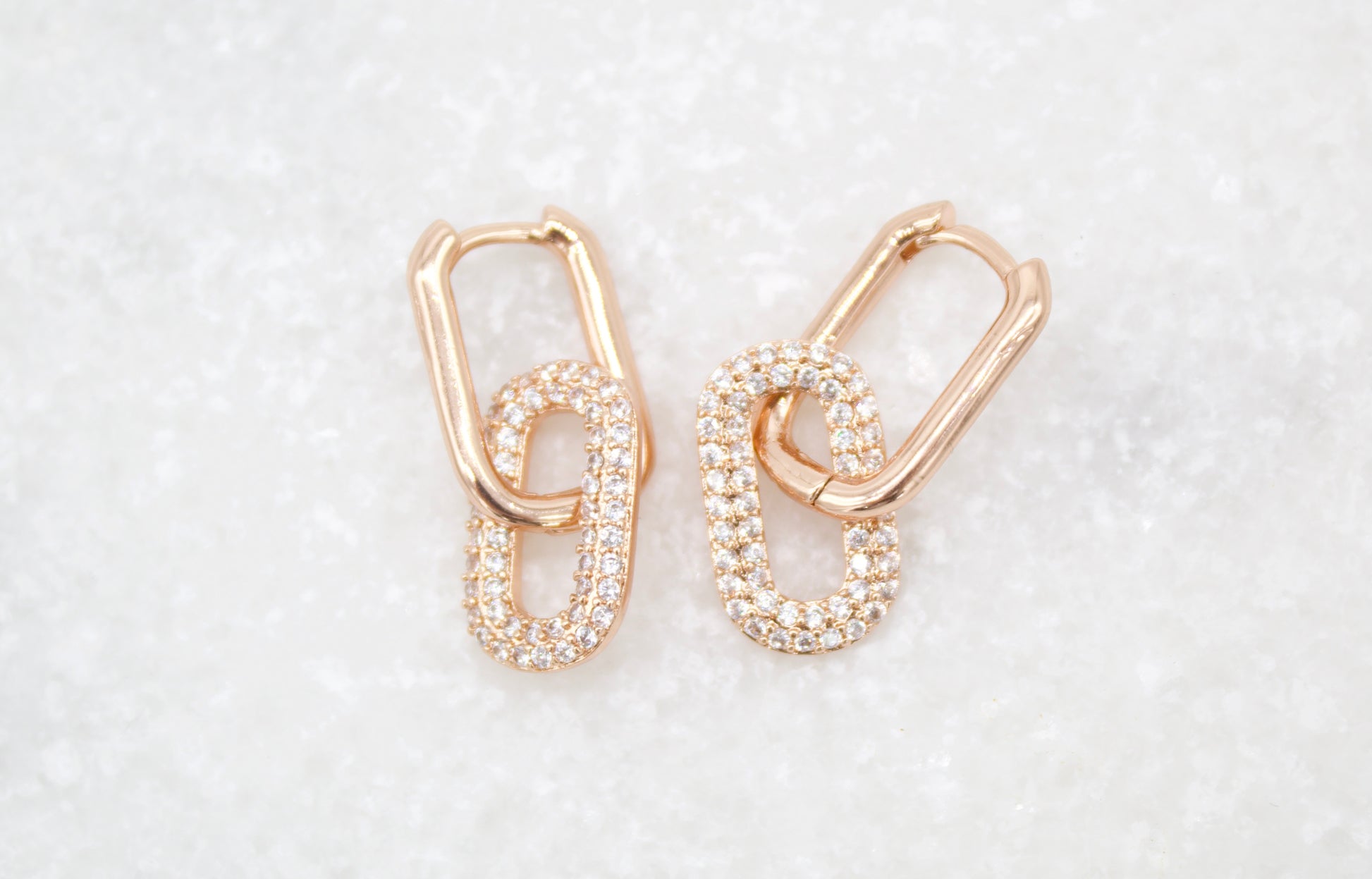 A pair of rose gold interlocking chain link earrings featuring pavé-set diamonds rests on a textured white surface.