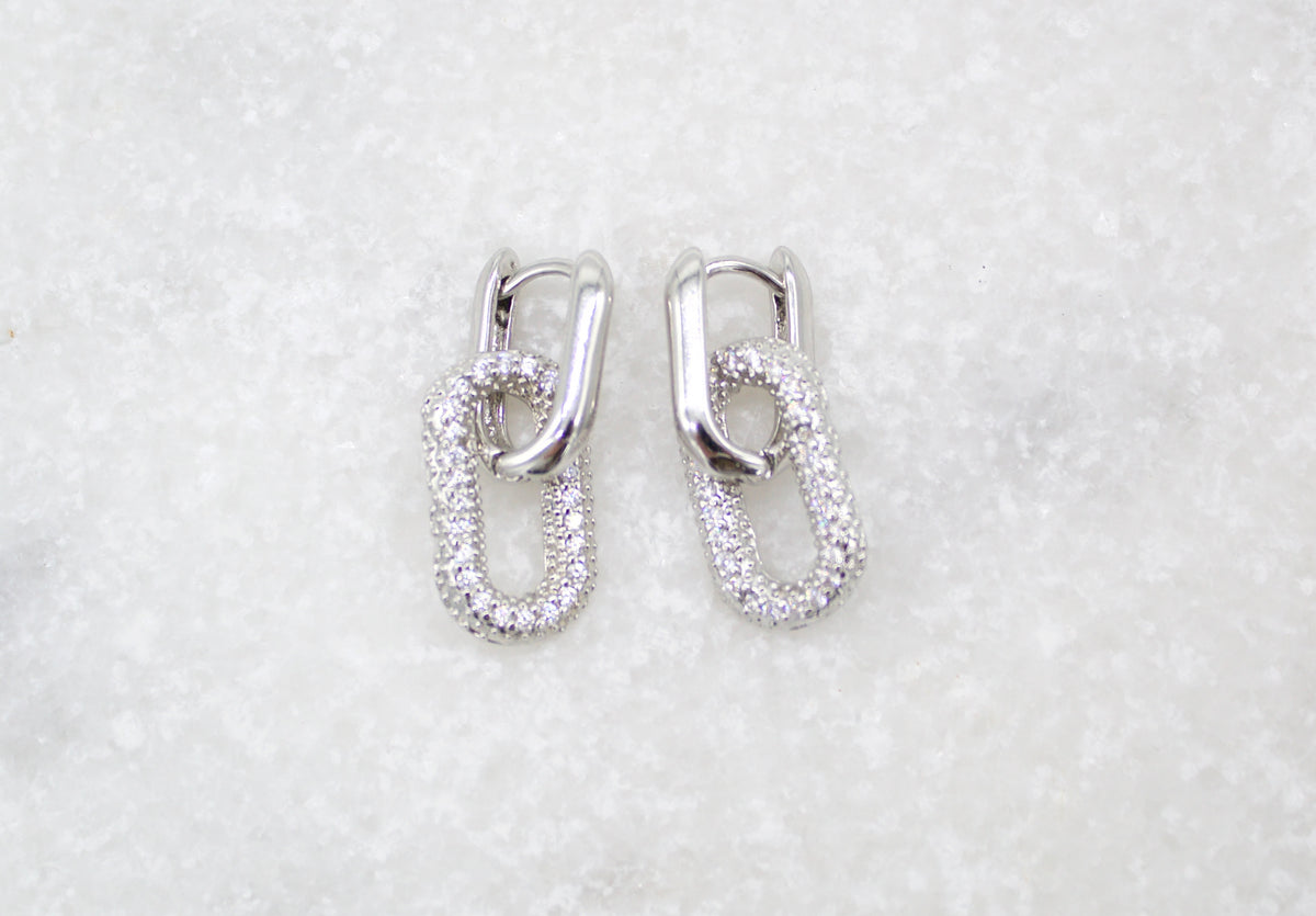 A pair of sparkling silver pave-set huggie hoop earrings featuring interlocking links rests against a white marble surface.
