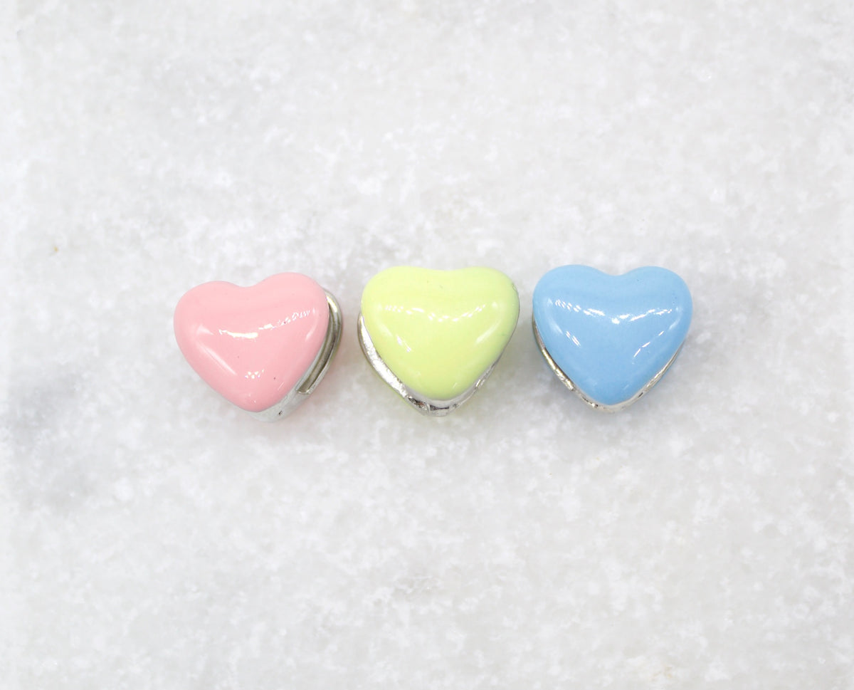 Three glossy pastel heart-shaped beads in pink, yellow, and blue lie in a row on a white surface.