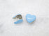 A pair of glossy light blue heart-shaped huggie hoop earrings rests on a textured white surface.