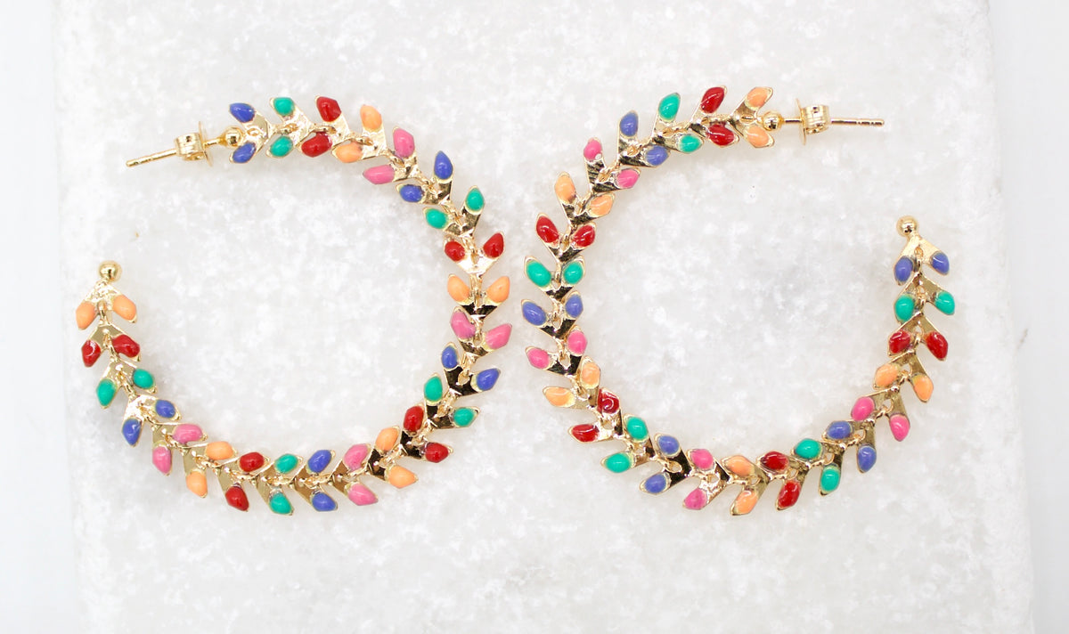 A pair of gold hoop earrings embellished with colorful enamel leaves rests on a textured white surface.
