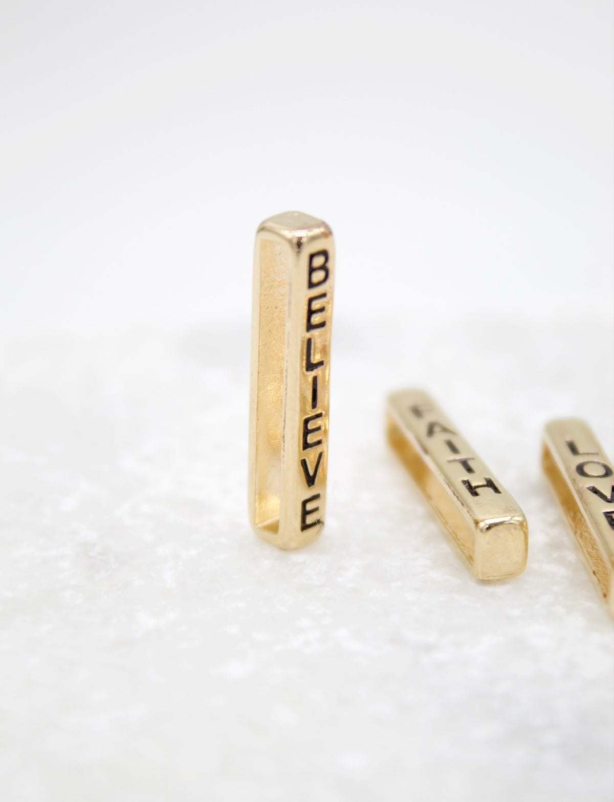 A vertical gold rectangular pendant with the word "BELIEVE" stamped on it rests on a white surface alongside two other similar pendants.