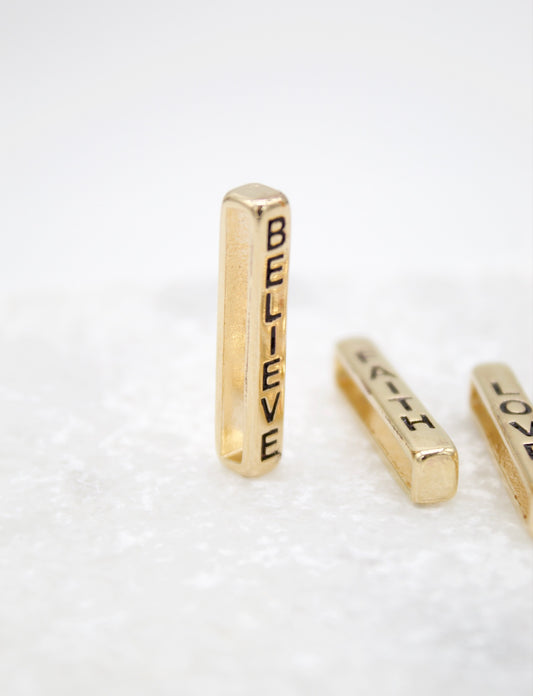 A vertical gold bar bead inscribed with the word "BELIEVE" stands upright on a white surface next to other word charms.