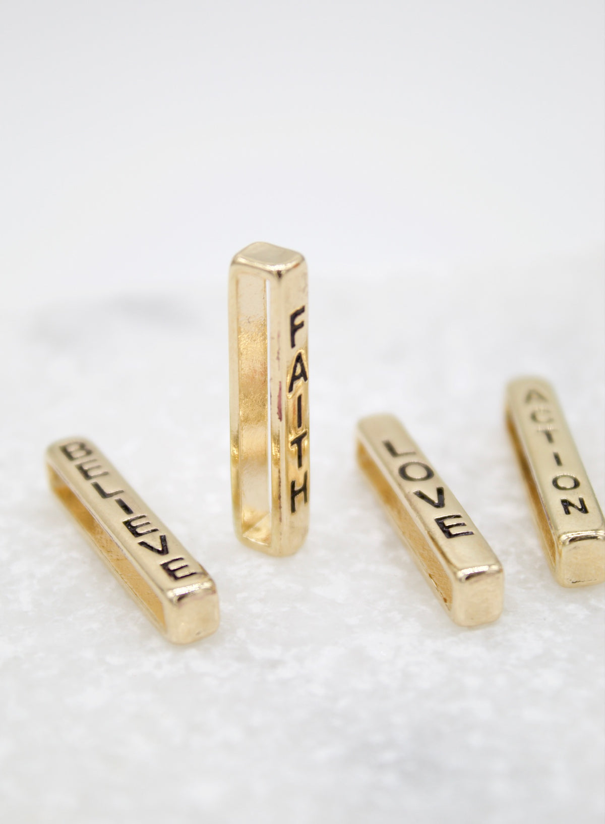 Four gold rectangular slider beads engraved with the words "BELIEVE," "FAITH," "LOVE," and "ACTION" stand upright on a white surface.