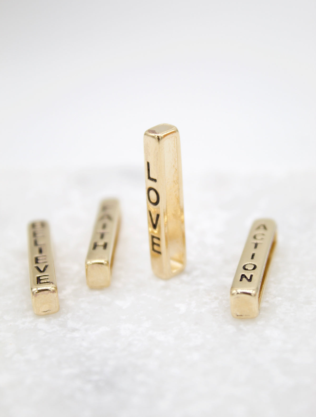 Four gold bar pendants engraved with the words "LOVE," "FAITH," "BELIEVE," and "ACTION" stand upright on a textured white surface.