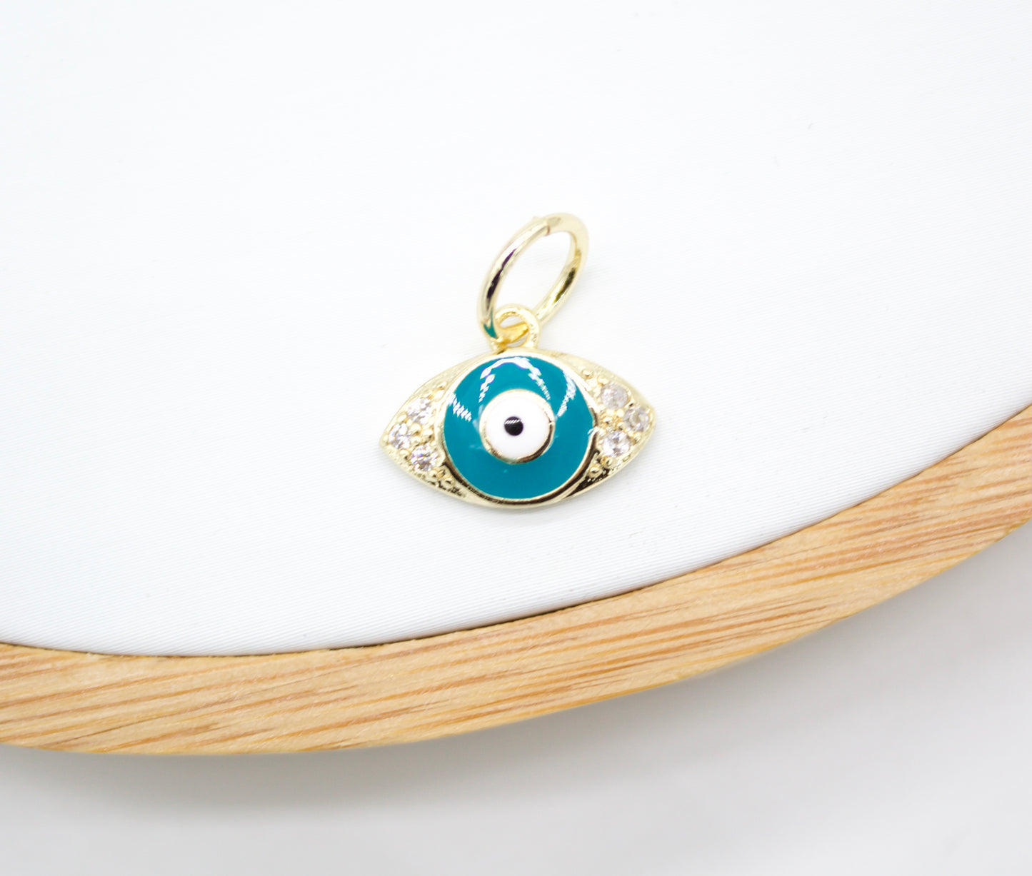 A gold evil eye charm pendant featuring blue enamel and sparkling cubic zirconia stones rests on a white display surface.