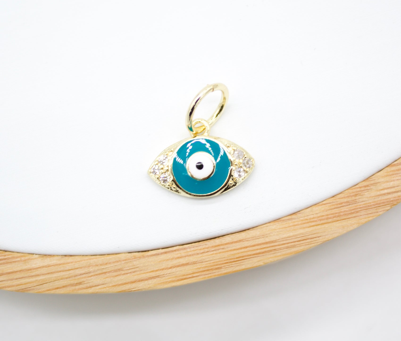 A gold evil eye charm pendant featuring blue enamel and sparkling cubic zirconia stones rests on a white display surface.