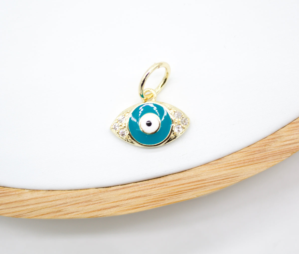A gold evil eye charm pendant featuring blue enamel and sparkling cubic zirconia stones rests on a white display surface.