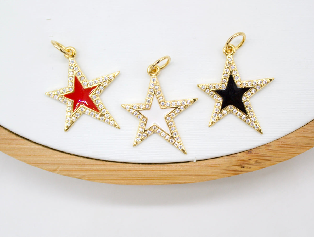 Three gold star pendant charms featuring pave cubic zirconia and colorful enamel in red, white, and black rest on a white surface with a wooden frame.