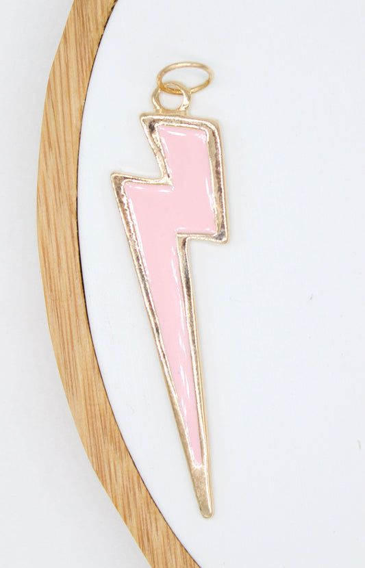 A gold-trimmed light pink lightning bolt enamel charm rests on a white display board inside a wooden embroidery hoop.