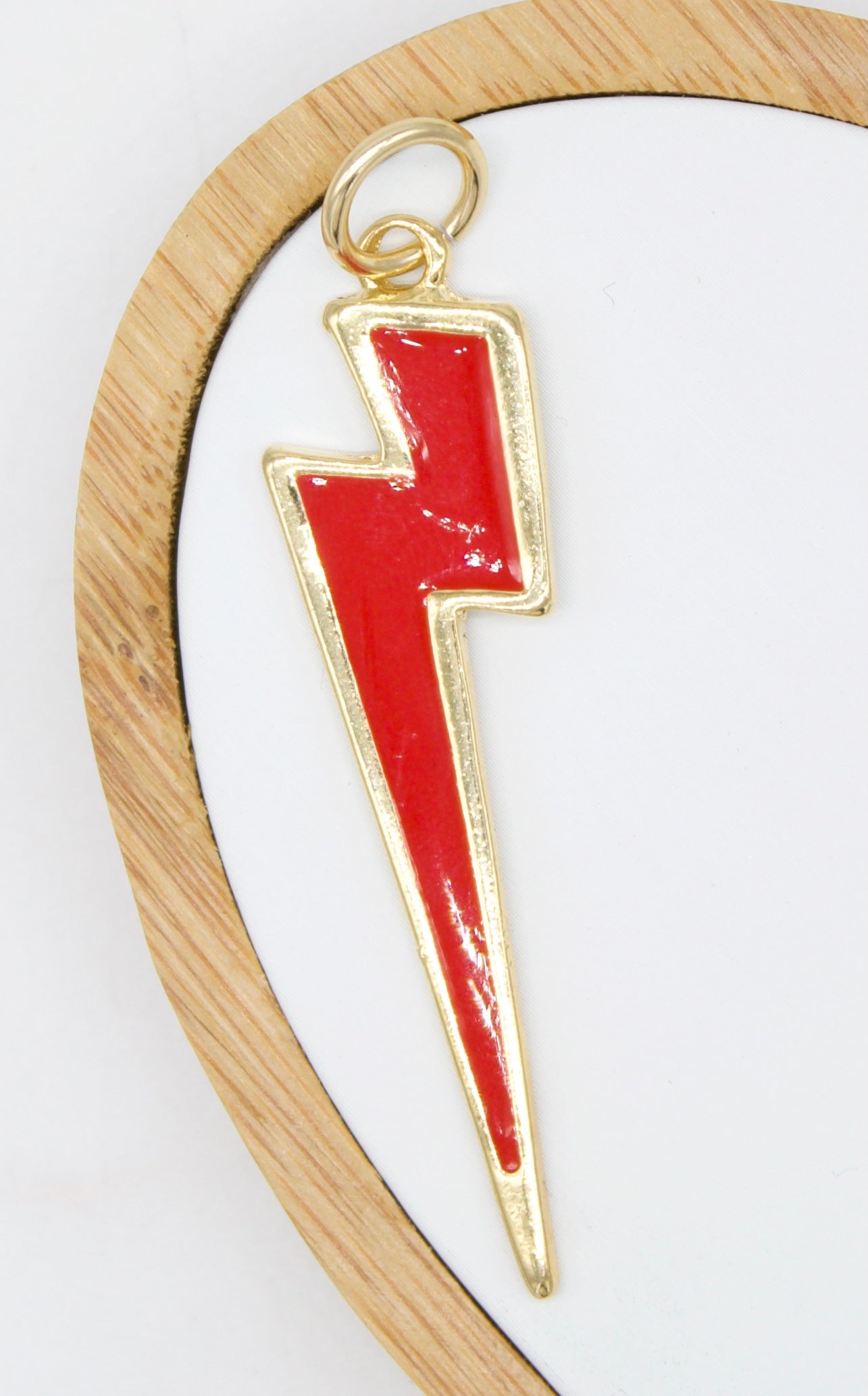 A shiny gold lightning bolt pendant features a vibrant red enamel inlay against a white background inside a round wooden frame.