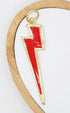 A shiny gold lightning bolt pendant features a vibrant red enamel inlay against a white background inside a round wooden frame.