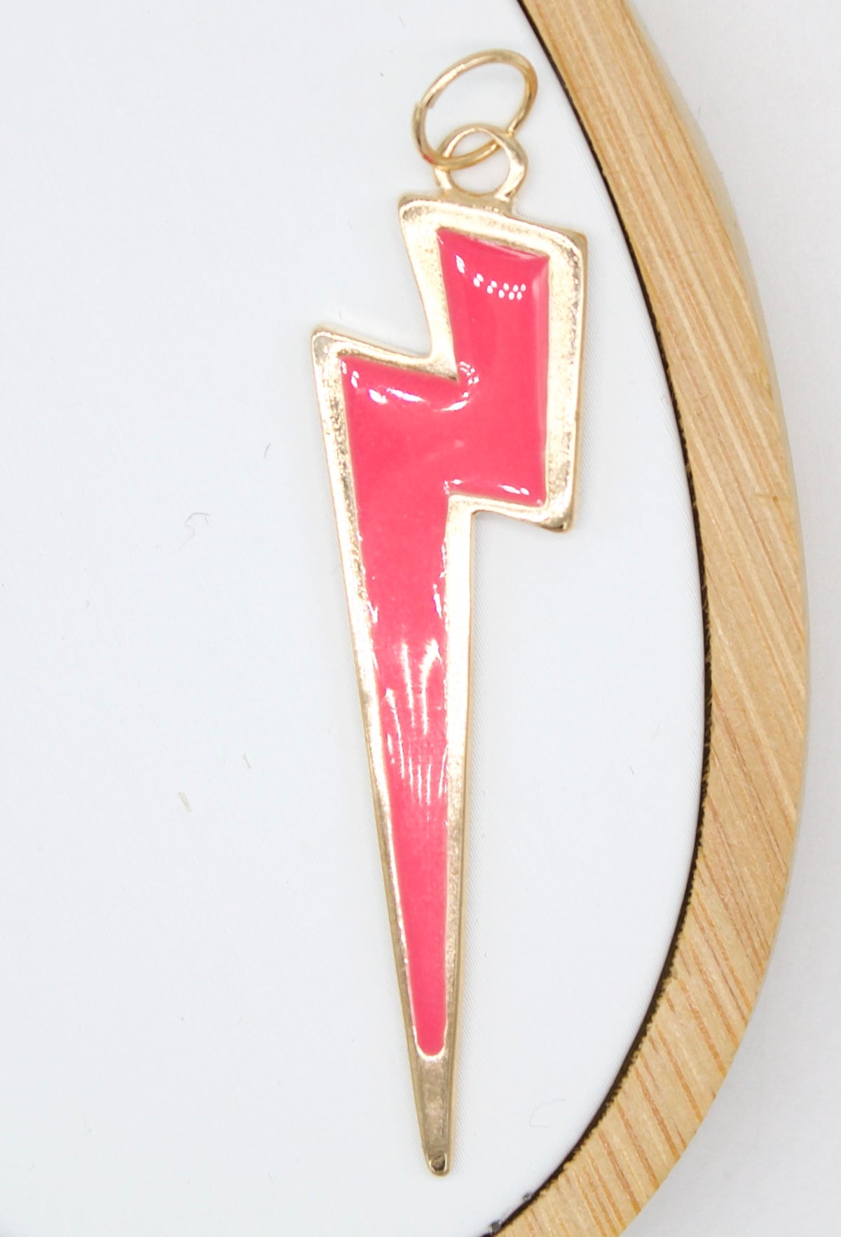 A gold-rimmed enamel pink lightning bolt charm pendant rests against a white fabric backdrop inside a round wooden embroidery hoop.