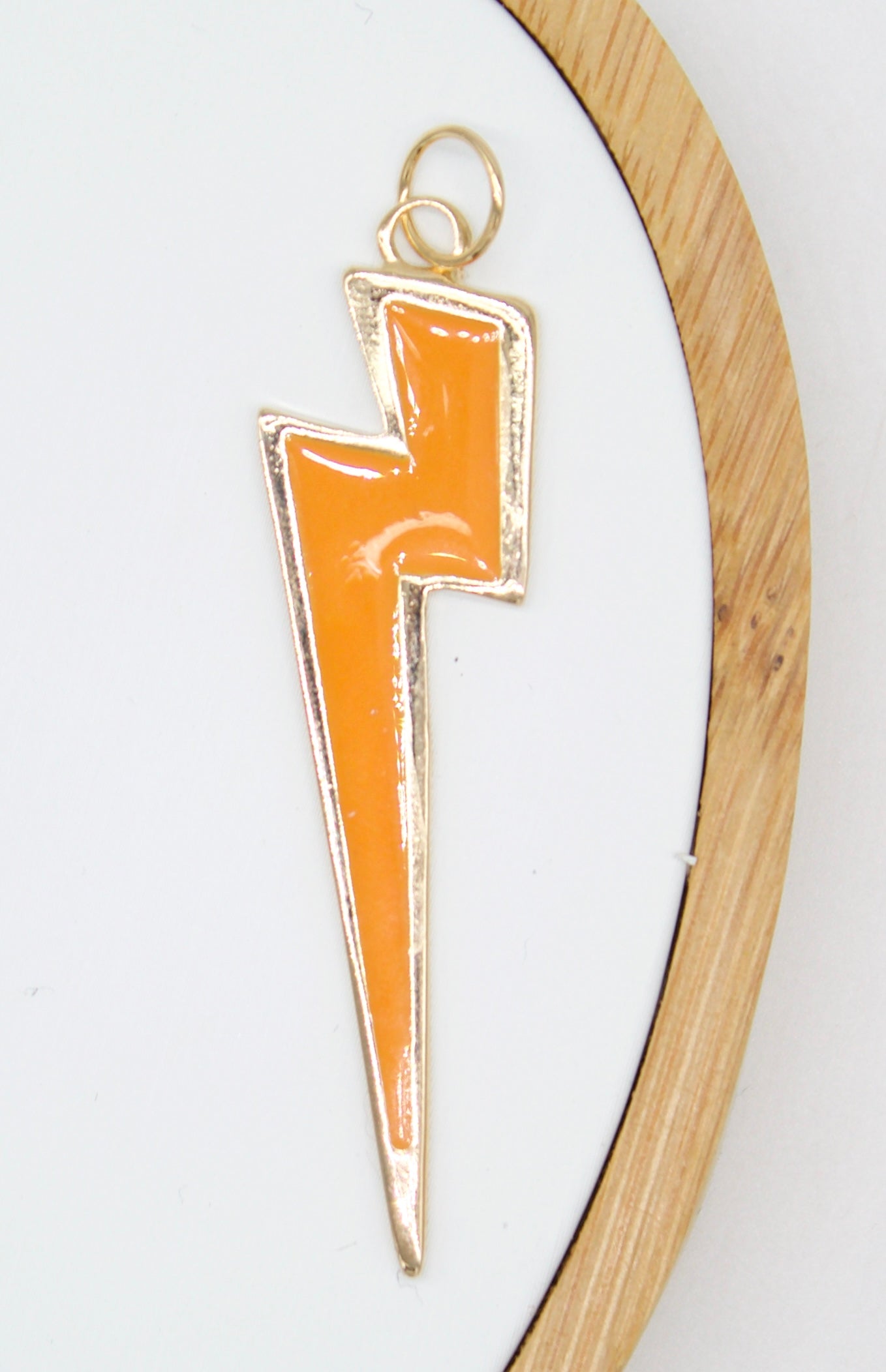 An orange enamel lightning bolt charm with gold trim rests against a white background inside a wooden embroidery hoop.