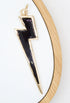 A gold-rimmed lightning bolt charm filled with glossy black enamel rests on a white display surface with a wooden frame.