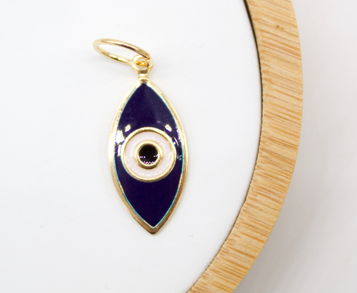 A gold evil eye pendant with dark blue and white enamel rests on a white display surface next to a wooden ring.