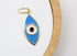 A gold-rimmed evil eye charm featuring blue and white enamel rests on a white display surface.