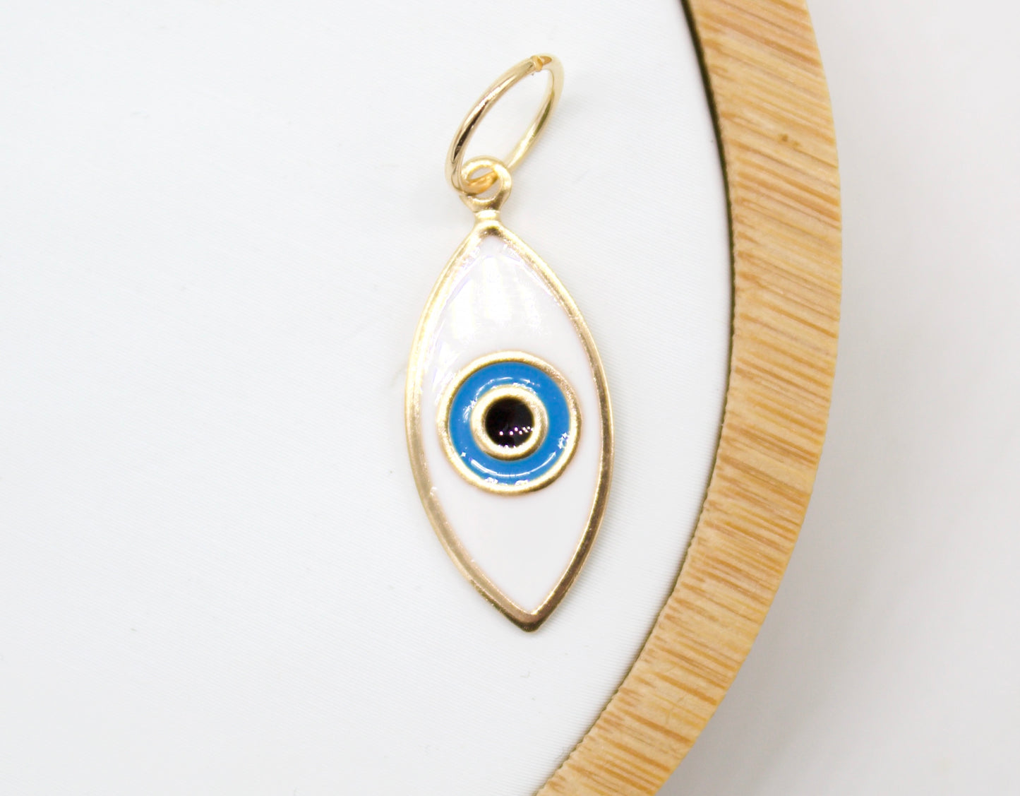 A delicate gold evil eye charm featuring white enamel and blue concentric circles rests on a neutral white display tray.