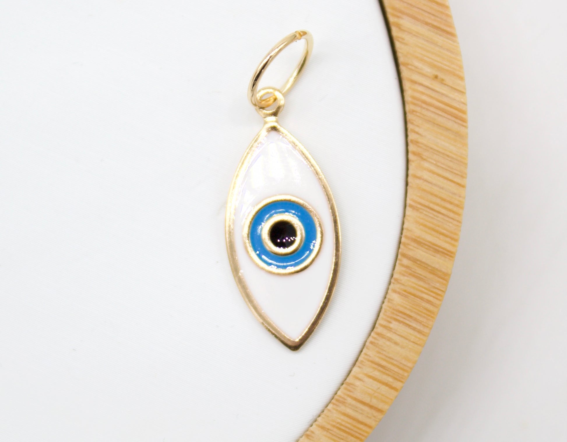 A delicate gold evil eye charm featuring white enamel and blue concentric circles rests on a neutral white display tray.