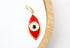 A gold evil eye pendant featuring red enamel rests on a white and wooden display surface.