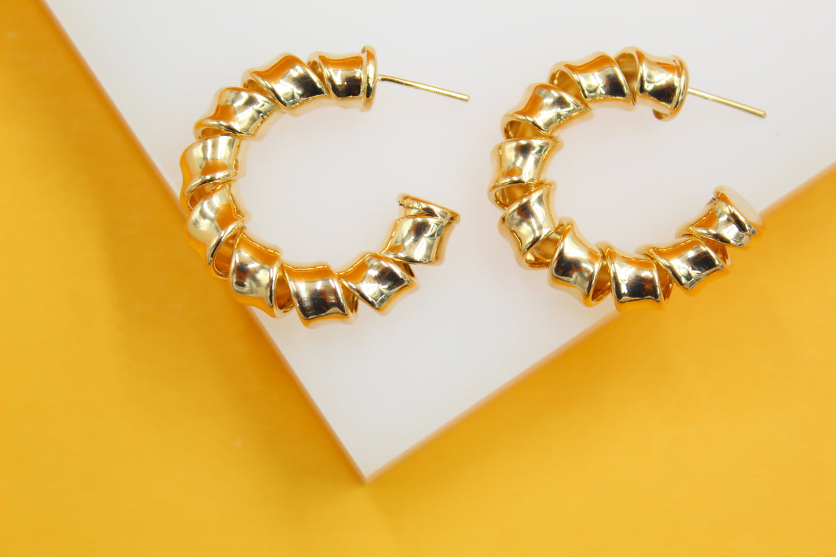 A pair of shiny gold twisted hoop earrings rests on a white and yellow display surface.