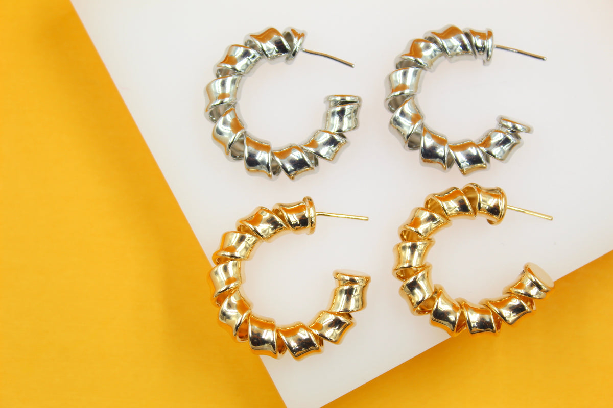 Two pairs of metallic spiral hoop earrings, one in silver and one in gold, rest on a white and yellow display background.
