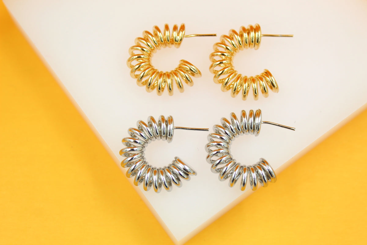 A pair of gold and a pair of silver chunky spiral hoop earrings rest on a white display stand against a vibrant yellow background.